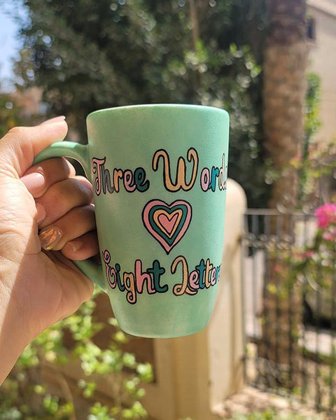 A Hand painted green Mug with the words "three words, Eight Letters" written and a heart place in between the two sentences