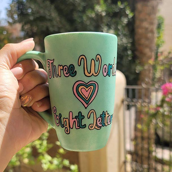 A Hand painted green Mug with the words "three words, Eight Letters" written and a heart place in between the two sentences