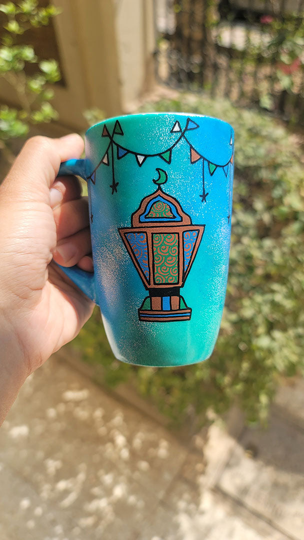 A Blue Green Hand painted mug with decorations hanging from the top all around the mug and a fanoos painted in black brown, blue and green