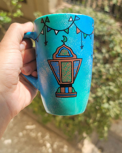 A Blue Green Hand painted mug with decorations hanging from the top all around the mug and a fanoos painted in black brown, blue and green