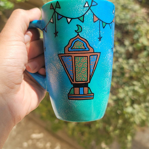 A Blue Green Hand painted mug with decorations hanging from the top all around the mug and a fanoos painted in black brown, blue and green