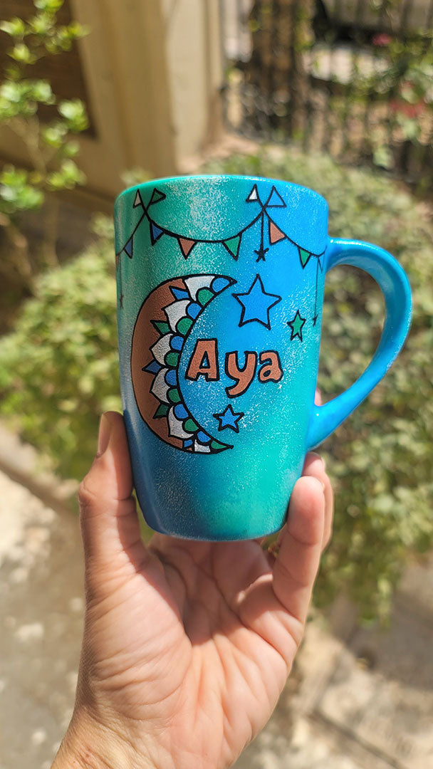 A Blue Green Hand painted mug with decorations hanging from the top all around the mug and a moon painted in black brown, blue and green and the name Aya written beside it