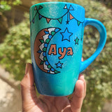 A Blue Green Hand painted mug with decorations hanging from the top all around the mug and a moon painted in black brown, blue and green and the name Aya written beside it