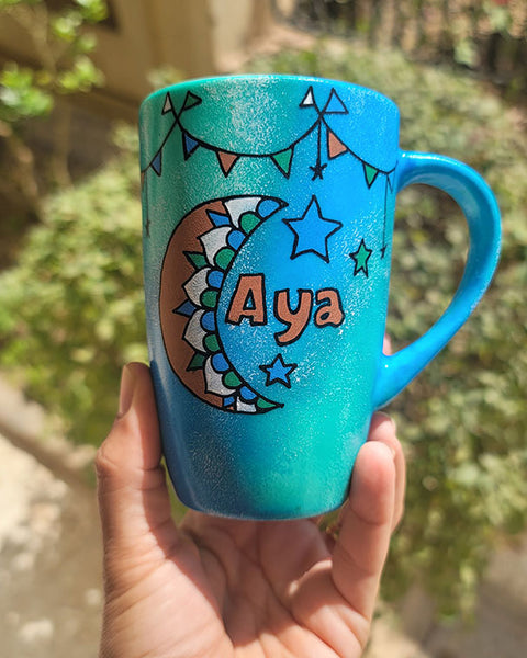 A Blue Green Hand painted mug with decorations hanging from the top all around the mug and a moon painted in black brown, blue and green and the name Aya written beside it