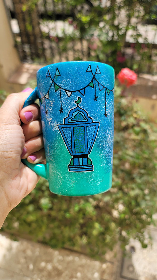 A Blue Green Hand painted mug with decorations hanging from the top all around the mug and a fanoos painted in black , blue and green