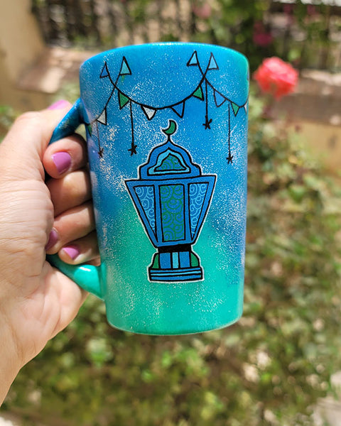 A Blue Green Hand painted mug with decorations hanging from the top all around the mug and a fanoos painted in black , blue and green