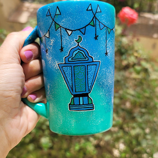 A Blue Green Hand painted mug with decorations hanging from the top all around the mug and a fanoos painted in black , blue and green