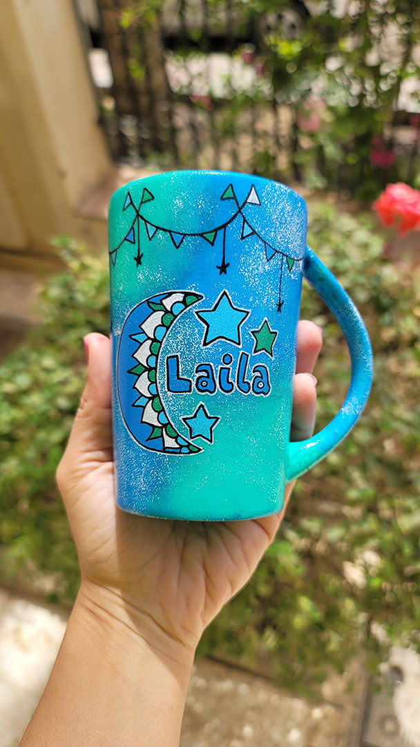 A Blue Green Hand painted mug with decorations hanging from the top all around the mug and a moon painted in black, blue and green and the name laila beside it