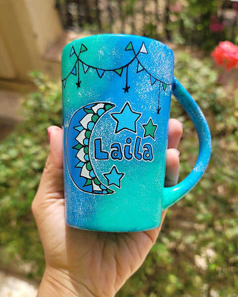 A Blue Green Hand painted mug with decorations hanging from the top all around the mug and a moon painted in black, blue and green and the name laila beside it