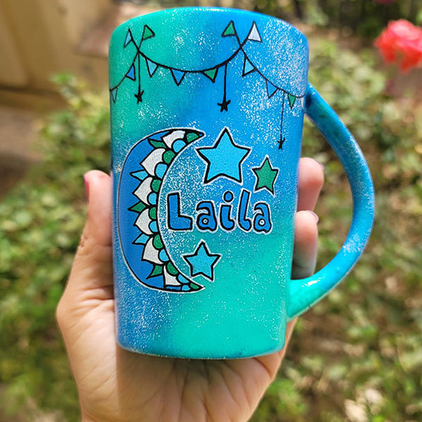 A Blue Green Hand painted mug with decorations hanging from the top all around the mug and a moon painted in black, blue and green and the name laila beside it