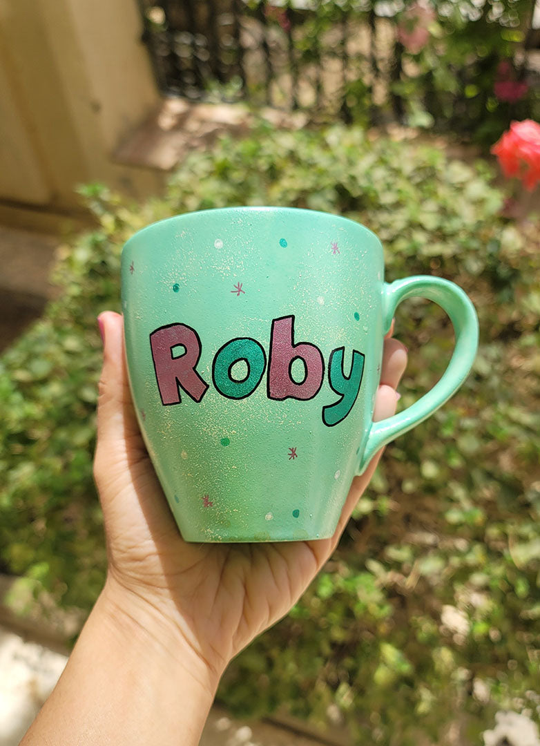 A mint Colored mug with the name "Roby" with white and green dots and pink stars scattered all over