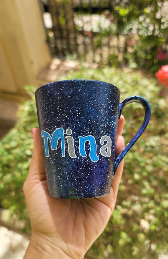 a Bluish galaxy hand painted mug with the name "mina"