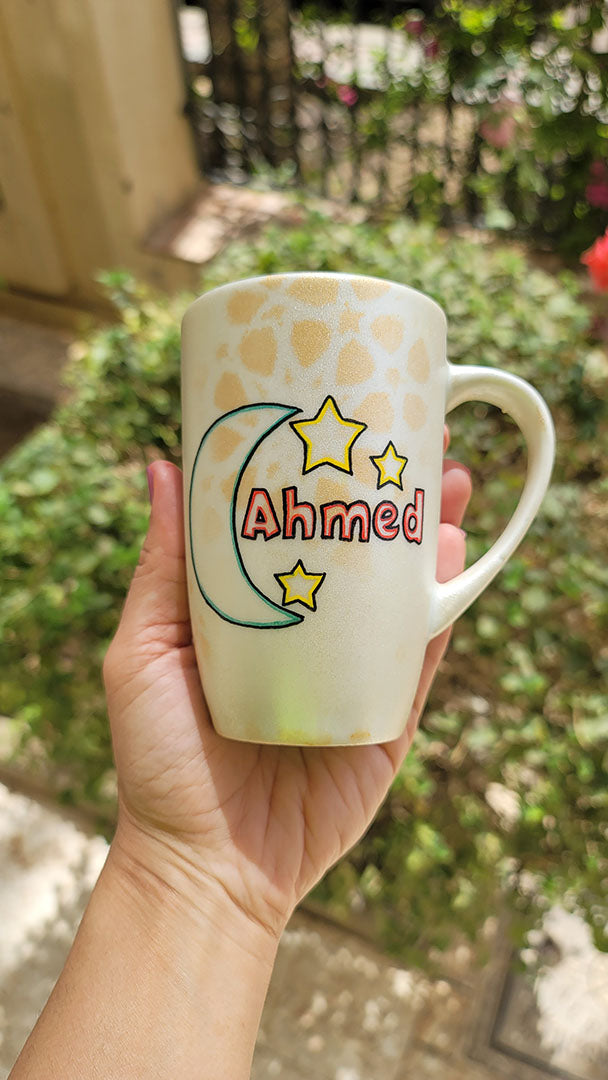 A Shimmery White with gold islamic patterns hand painted mug with a moon painted in black and blue green and the name Ahmed beside it