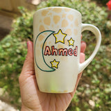 A Shimmery White with gold islamic patterns hand painted mug with a moon painted in black and blue green and the name Ahmed beside it