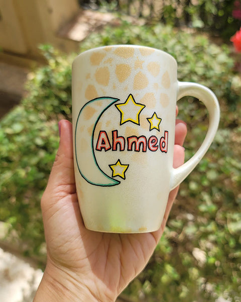 A Shimmery White with gold islamic patterns hand painted mug with a moon painted in black and blue green and the name Ahmed beside it