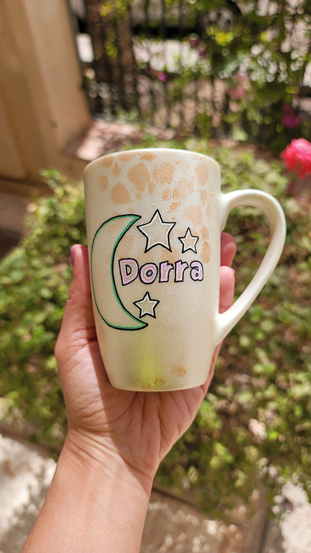 A Shimmery White with gold islamic patterns hand painted mug with a moon painted in black and blue green and the name Dorra beside it