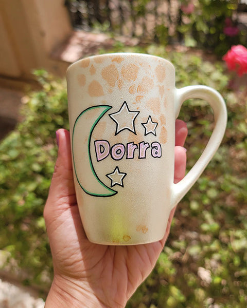 A Shimmery White with gold islamic patterns hand painted mug with a moon painted in black and blue green and the name Dorra beside it