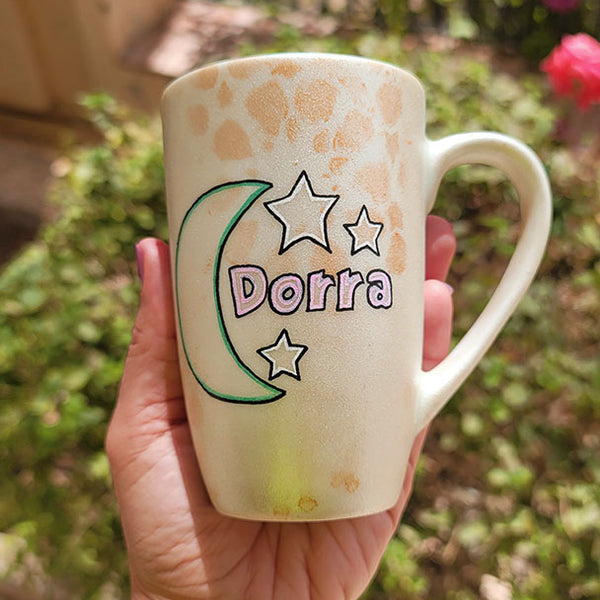 A Shimmery White with gold islamic patterns hand painted mug with a moon painted in black and blue green and the name Dorra beside it