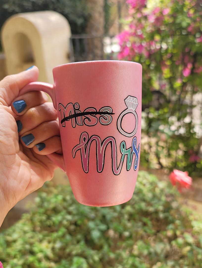 A shimmery pink hand-painted mug with the word miss crossed out and the words mrs in larger font, with a diamond ring drawn on the side