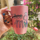 A shimmery pink hand-painted mug with the word miss crossed out and the words mrs in larger font, with a diamond ring drawn on the side