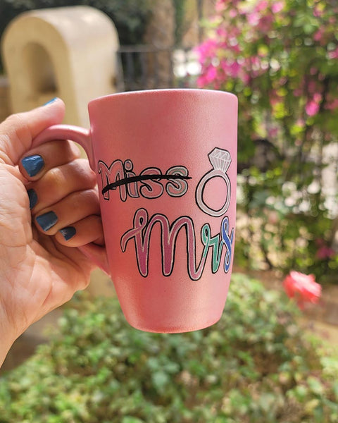 A shimmery pink hand-painted mug with the word miss crossed out and the words mrs in larger font, with a diamond ring drawn on the side