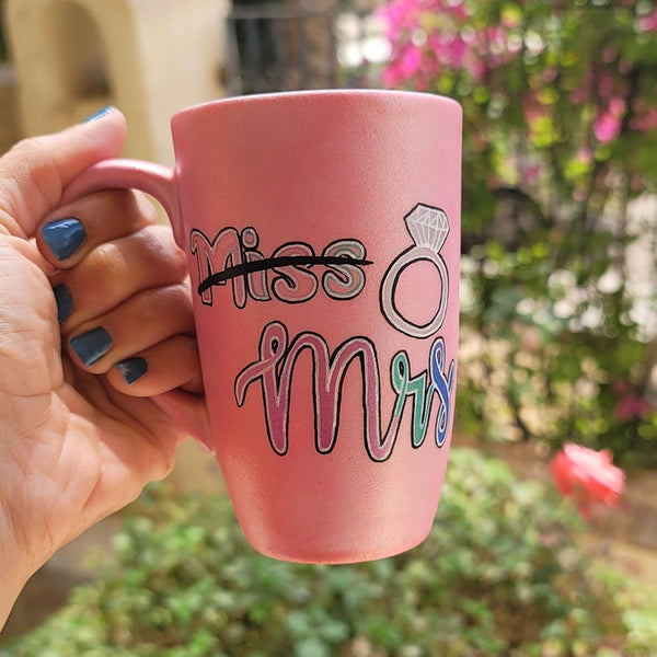 A shimmery pink hand-painted mug with the word miss crossed out and the words mrs in larger font, with a diamond ring drawn on the side