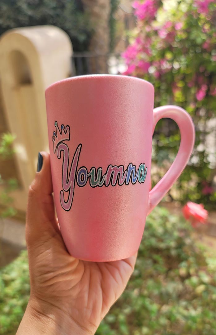 A shimmery pink hand-painted mug with the word miss crossed out and the name "Youmna" and a little crown on the letter Y