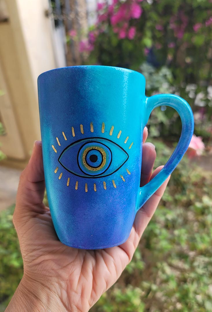 A hand painted blue mug with an eye painted in the center with gold eye lashes