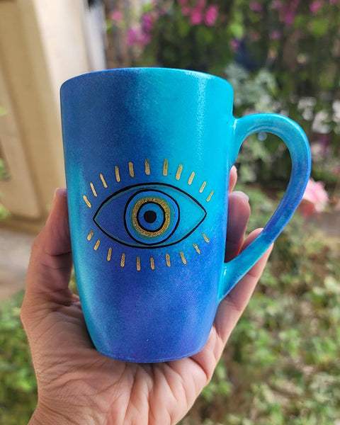 A hand painted blue mug with an eye painted in the center with gold eye lashes