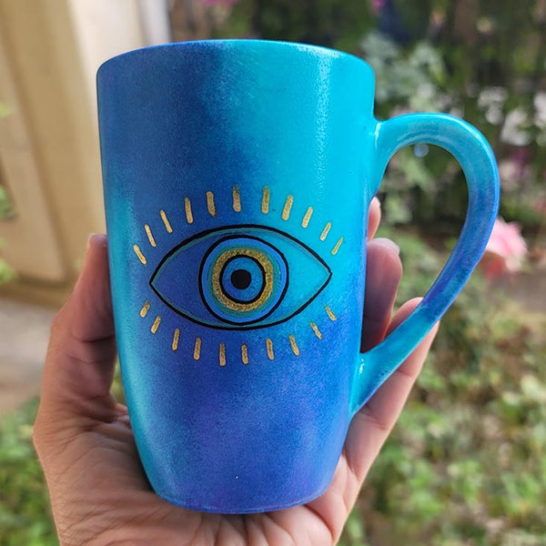 A hand painted blue mug with an eye painted in the center with gold eye lashes