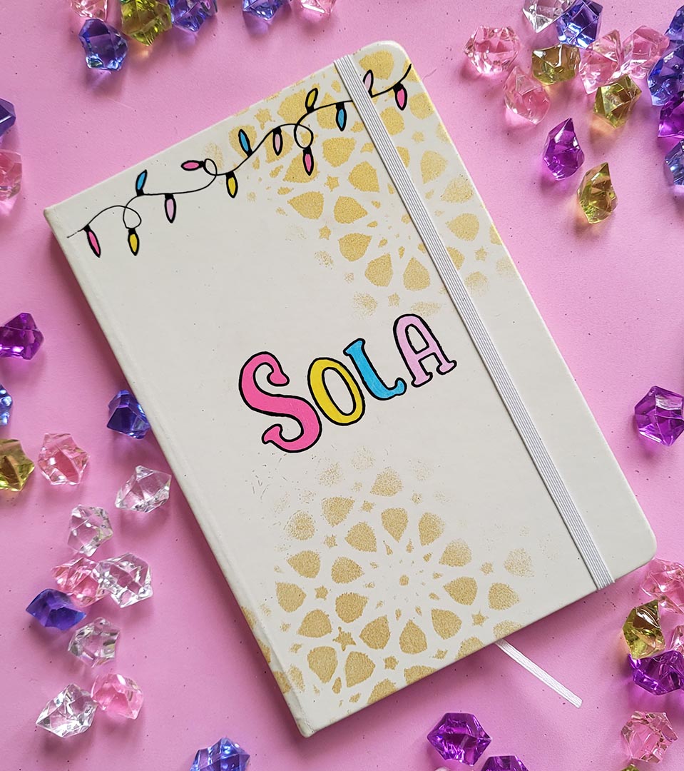 A white notebook with geometric patterns in gold in the background and festive lights all over the top, with the name "Sola" written in the center