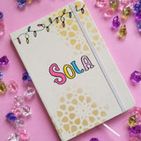 A white notebook with geometric patterns in gold in the background and festive lights all over the top, with the name 