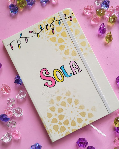 A white notebook with geometric patterns in gold in the background and festive lights all over the top, with the name "Sola" written in the center