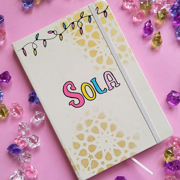 A white notebook with geometric patterns in gold in the background and festive lights all over the top, with the name "Sola" written in the center