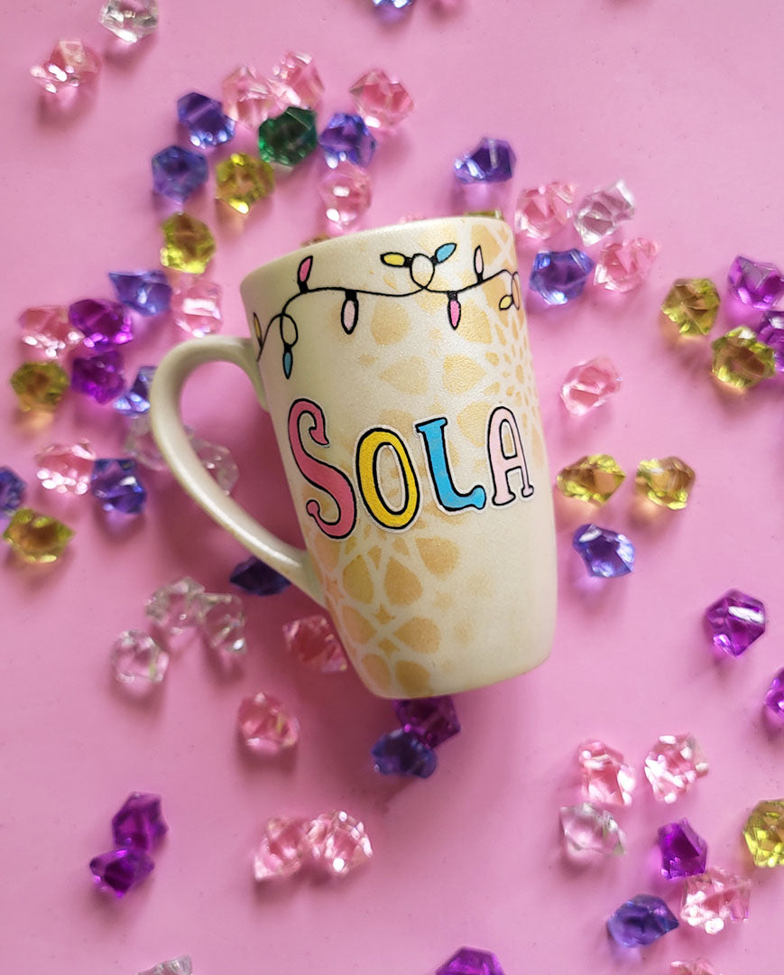 A Pearl white hand painted mug with the name sola on it and geometric  patterns in the background in gold with birthday lighting all over the top of the mug