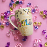 A Pearl white hand painted mug with the name sola on it and geometric  patterns in the background in gold with birthday lighting all over the top of the mug