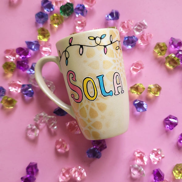 A Pearl white hand painted mug with the name sola on it and geometric  patterns in the background in gold with birthday lighting all over the top of the mug