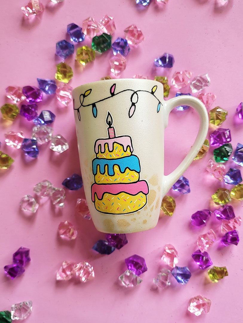 A Pearl white hand painted mug with a layered cake painted  in yellow and icing of baby pink , blue and fushia, with sprinkles all over the yellow cake and a pink candle on top, also the mug has birthday lighting all over the top of the mug