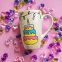 A Pearl white hand painted mug with a layered cake painted  in yellow and icing of baby pink , blue and fushia, with sprinkles all over the yellow cake and a pink candle on top, also the mug has birthday lighting all over the top of the mug