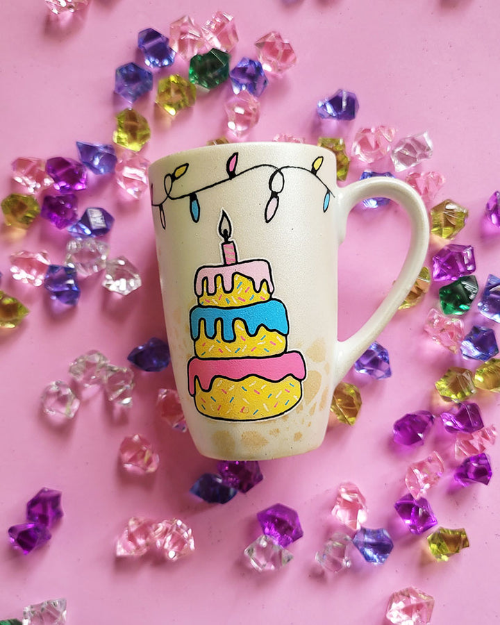 A Pearl white hand painted mug with a layered cake painted  in yellow and icing of baby pink , blue and fushia, with sprinkles all over the yellow cake and a pink candle on top, also the mug has birthday lighting all over the top of the mug