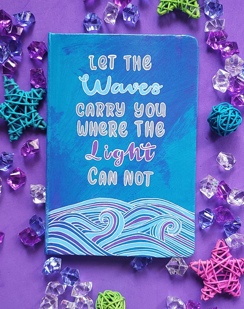 A Blue Hand painted notebook with patterns of waves drawn in the bottom and the words "let the waves carry you where the light can not"