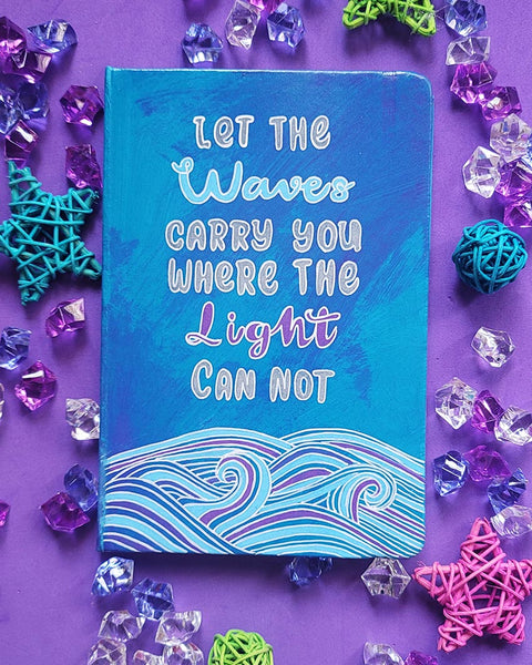 A Blue Hand painted notebook with patterns of waves drawn in the bottom and the words "let the waves carry you where the light can not"