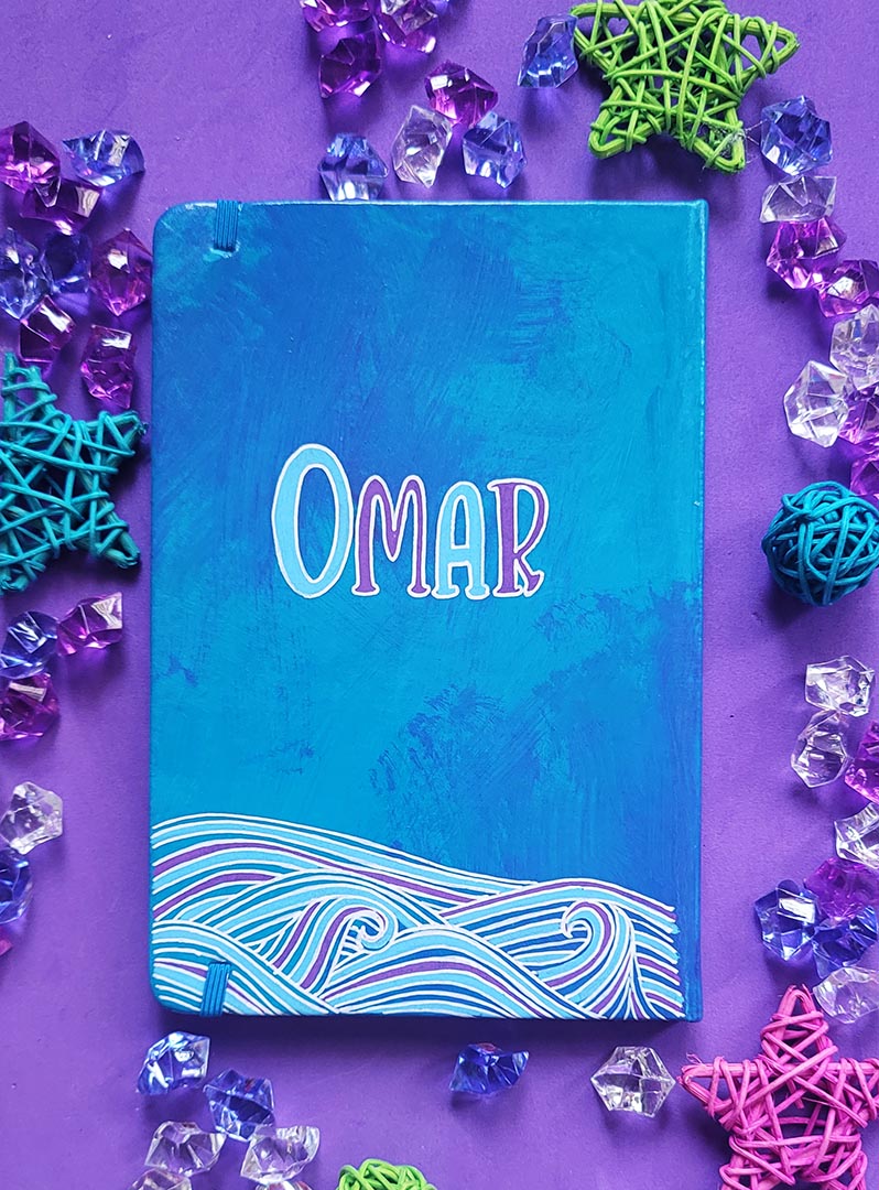 A Blue Hand painted notebook with patterns of waves drawn in the bottom and the name "Omar""