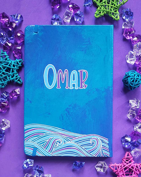 A Blue Hand painted notebook with patterns of waves drawn in the bottom and the name "Omar""