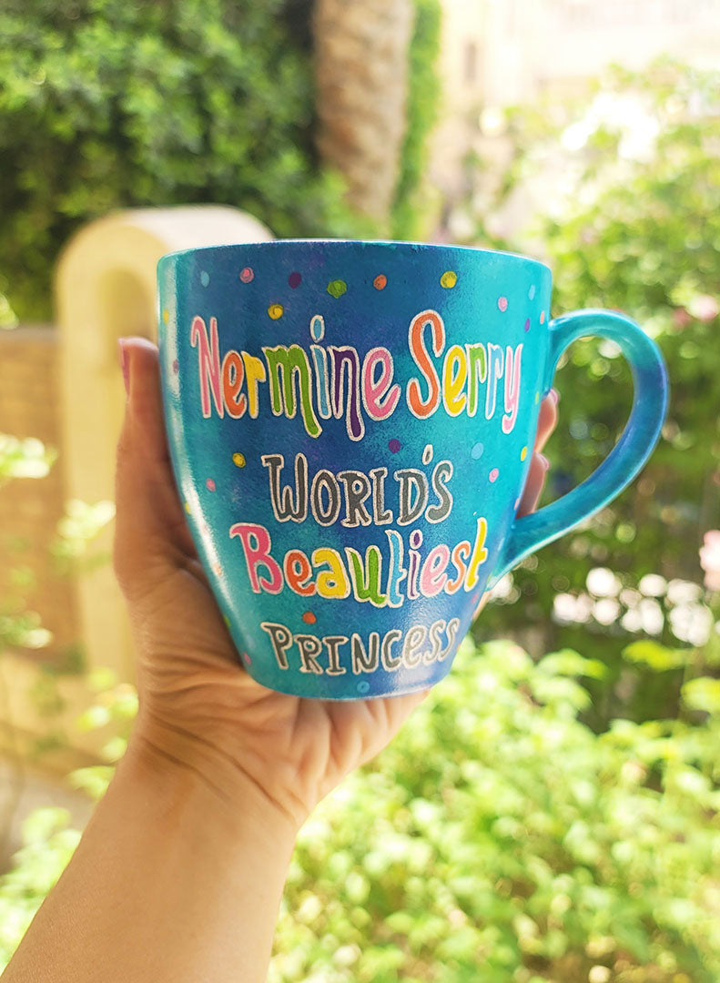 A blue hand painted mug with the words "Nermine Serry, worlds beautiest princess" and colorful dots scattered all over