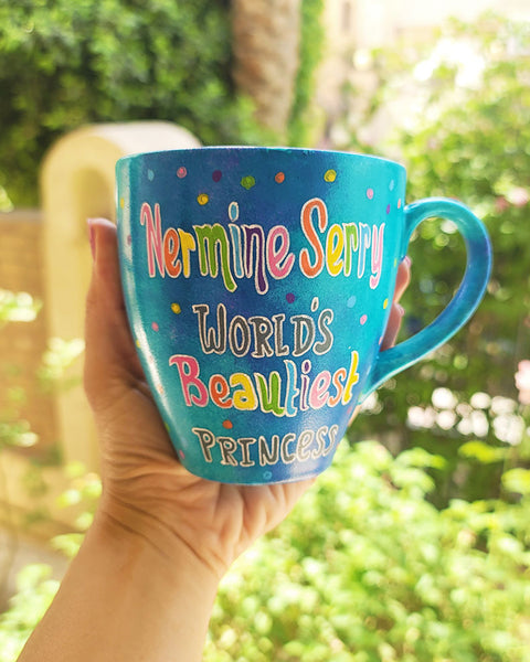 A blue hand painted mug with the words "Nermine Serry, worlds beautiest princess" and colorful dots scattered all over