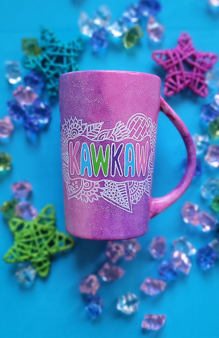 A pink galaxy hand painted mug with the name "kawkaw" inside a cloud of doodles