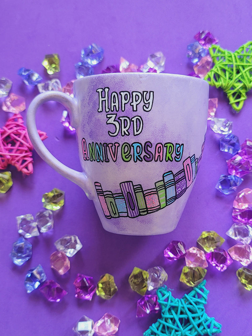 A Lilac hand painted mug with the words " happy 3rd anniversary" and book shelves rising diagonally on the mug