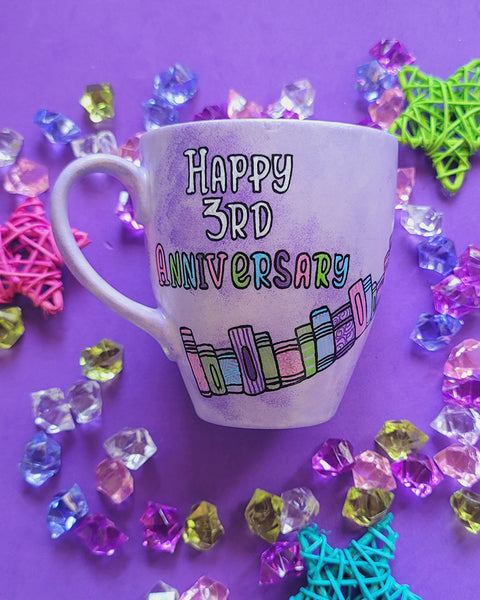 A Lilac hand painted mug with the words " happy 3rd anniversary" and book shelves rising diagonally on the mug