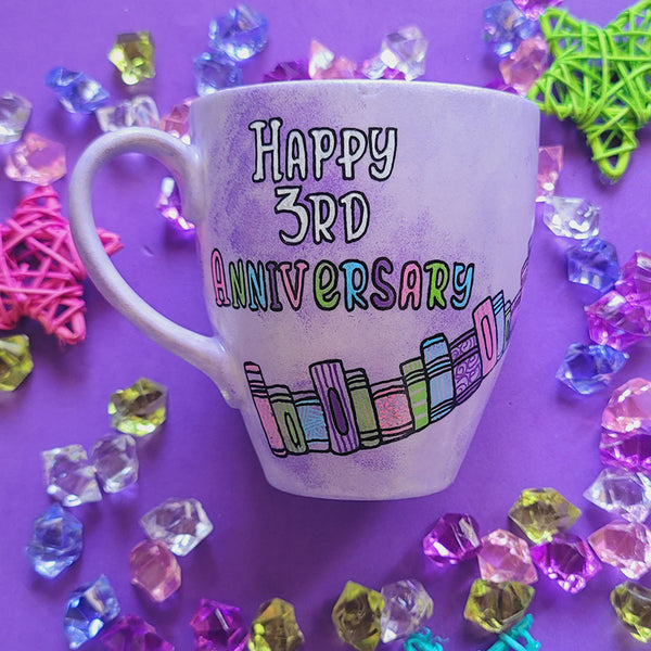 A Lilac hand painted mug with the words " happy 3rd anniversary" and book shelves rising diagonally on the mug